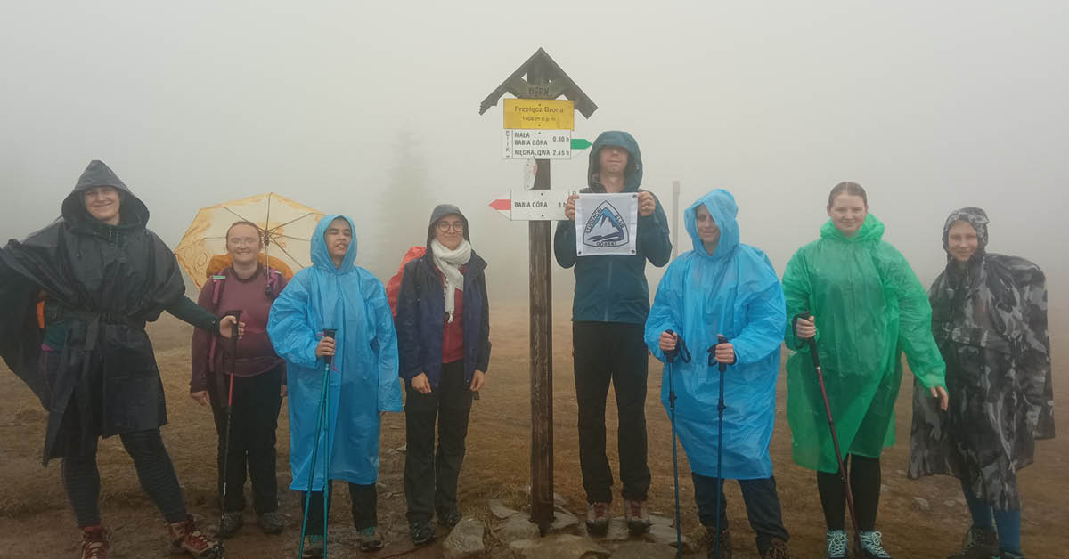 Grupa ludzi z flagą SKG, wszyscy ubrani w kurtki i płaszcze przeciwdeszczowe, za nimi widać tylko mgłę. 